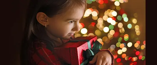 Enfant à Noël tenant un cadeau représentant une carte à gratter près du sapin illuminé
